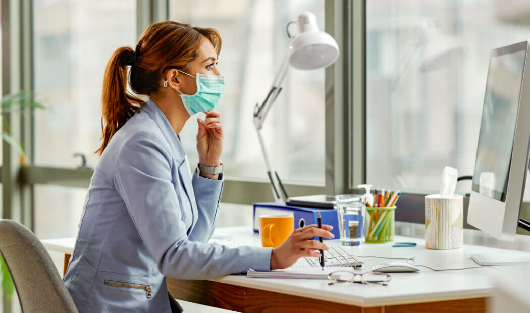worker wearing a face mask sat at a desk in front of monitor